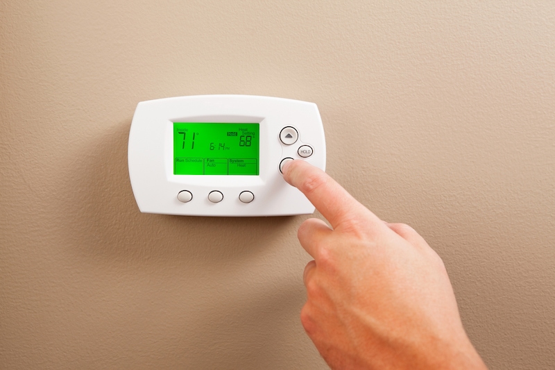 As a energy saving measure a male hand is turning down a digital programmable thermostat. The temperature reads 71 degrees